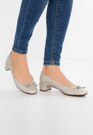 Pieds portant des chaussures en cuir verni beige clair à talon bas bloc avec détail nœud, associées à un jean skinny bleu.