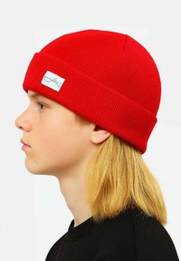 Red knitted beanie with a folded cuff, featuring a white logo patch. Soft texture and close-fitting design. No additional embellishments.