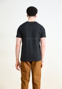 Musta puuvillapaita, jossa on tekstuurillinen takapainatus "ALPHA INDUSTRIES" ja pyöreä kaula-aukko. Yhdistetty vaaleanruskeisiin housuihin.