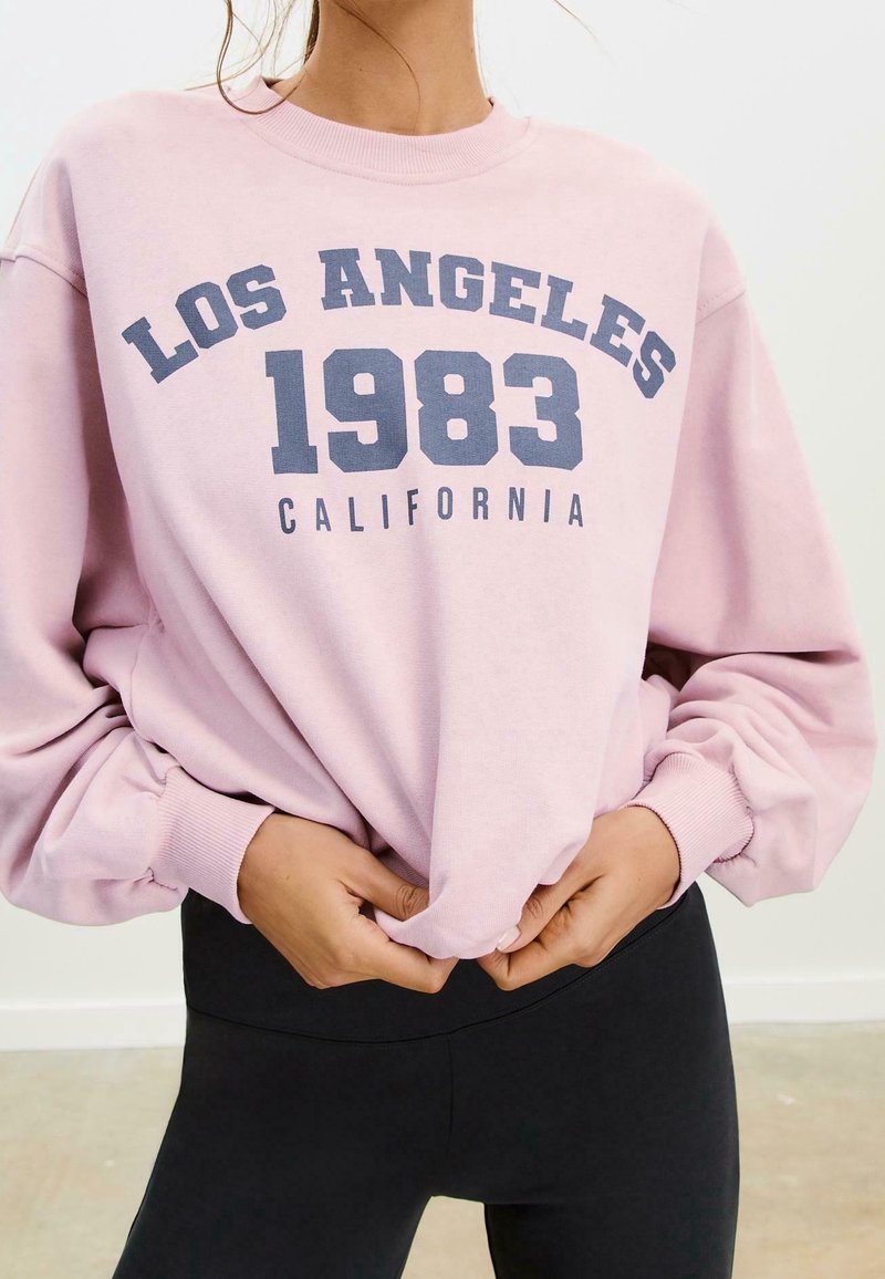 Personne portant un sweat-shirt rose avec le texte "Los Angeles 1983 California", tenant l'ourlet inférieur, associé à un pantalon noir.