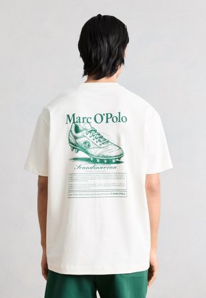 Osoba koja nosi bijelu majicu s zelenom ilustracijom nogometne kopačke i natpisom "Marc O'Polo" na leđima, u kombinaciji sa zelenim kratkim hlačama.