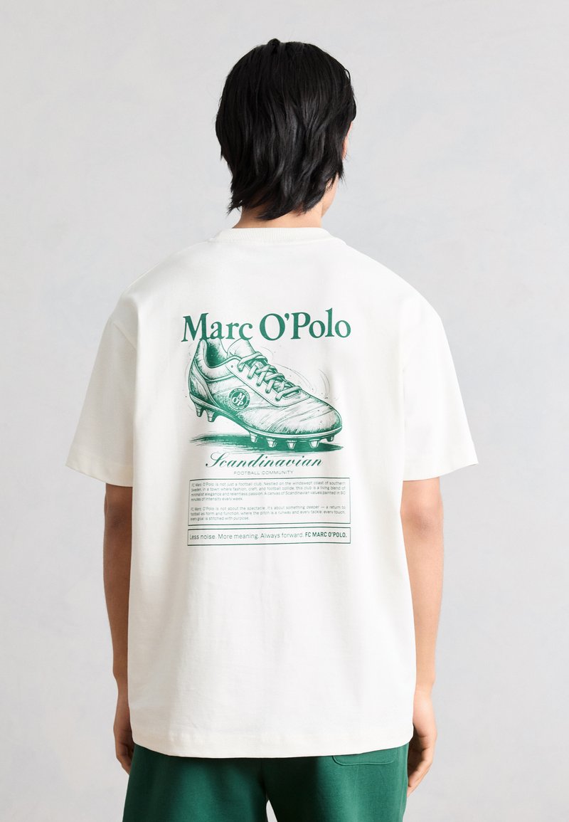 Άτομο που φοράει λευκό μπλουζάκι με πράσινη απεικόνιση ποδοσφαιρικού παπουτσιού και το κείμενο «Marc O'Polo» στην πλάτη, συνδυασμένο με πράσινα σορτς.