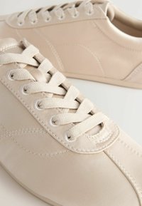 Beige sneakers made of smooth synthetic material, featuring a classic lace-up design, round toe, and textured eyelets.
