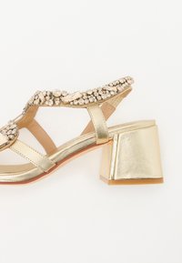 Gullfargede sandaler med klumpete hæl, pyntet med klare og lyse gullstein, med metalliske stropper og en strukturert såle.