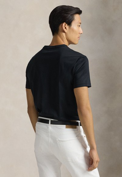 Άνδρας σε προφίλ φοράει μαύρο t-shirt, άσπρο τζιν και μαύρη ζώνη μπροστά σε ουδέτερο μπεζ φόντο.