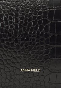 Portefeuille noir en cuir embossé crocodile avec une texture lisse et le logo doré "ANNA FIELD". Présente un design structuré et une forme rectangulaire.