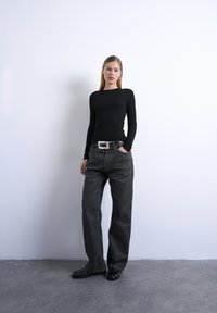 Jeune femme debout contre un mur blanc, portant un haut noir à manches longues, un pantalon ample gris foncé avec une ceinture blanche, et des chaussures noires.