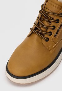 Chaussure en cuir brun avec une texture lisse, lacets noirs, semelle contrastante blanche et noire, et un détail de fermeture éclair sur le côté.