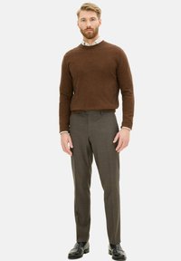 Maglione lavorato a maglia marrone con scollatura rotonda, abbinato a pantaloni a quadretti grigi chiari. Il modello indossa scarpe in pelle nera. Sfondo bianco semplice.