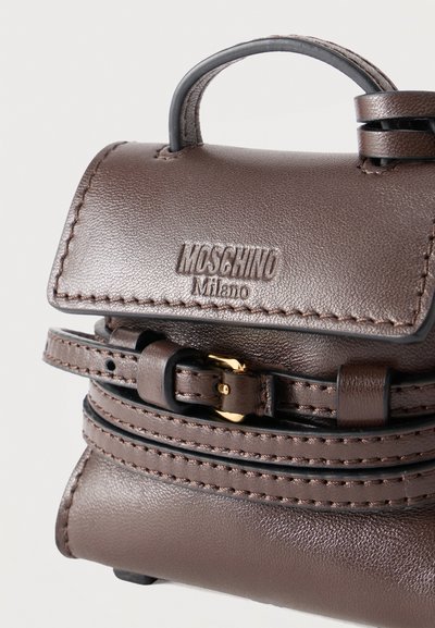 Μικρή καφέ δερμάτινη τσάντα χειρός Moschino Milano με πάνω χερούλι, λεπτομέρεια ζώνης και χρυσή αγκράφα σε απλό φόντο.