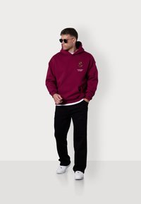 Bordeauxfarbener Kapuzenpullover mit floralem Design, Fronttasche, getragen mit schwarzen Hosen und weißen Sneakers, ergänzt durch eine dunkle Sonnenbrille.