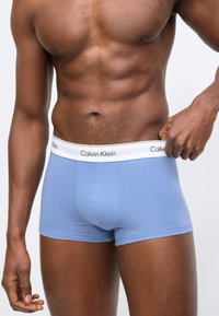 Μυώδης άνδρας που τραβά τη ζώνη μέσης απαλού γαλάζιου εσώρουχου Calvin Klein με λευκή ελαστική ταινία με το λογότυπο.