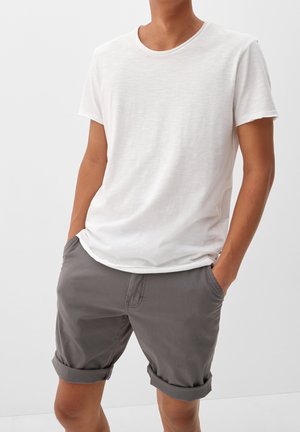 Mężczyzna w gładkiej, białej koszulce z krótkim rękawem i podwiniętych szarych szortach z rękami w kieszeniach na jednolitym tle.