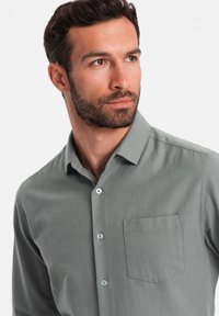 Chemise boutonnière grise claire en tissu doux, comportant une poche sur la poitrine et de petits boutons de couleur claire. Texture lisse et coupe ajustée.