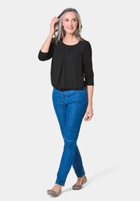 Schwarzes Oberteil mit Dreiviertelärmeln und Faltendetail, kombiniert mit schmal geschnittenen blauen Jeans und flachen grauen Schuhen, vor hellem Hintergrund.