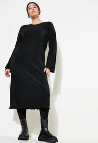Zwart geribbeld lang jurk met lange mouwen, golvende zoom, gedragen over zwarte leggings en gecombineerd met chunky zwarte enkellaarsjes.