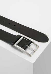 Cinturón de cuero negro con una hebilla de metal plateado, con múltiples agujeros de ajuste y una textura suave. Acentos: logotipo en la hebilla.