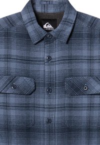 Camisa de franela a cuadros en azul marino y gris, con cuello, dos bolsillos en el pecho con cierres de botón y un forro suave y texturizado.