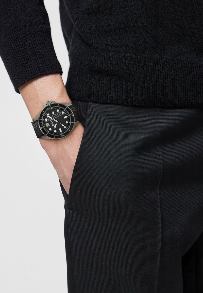 Person trägt schwarzen Pullover und Hose, hat die linke Hand in der Tasche und zeigt eine schwarze Armbanduhr mit gemustertem Armband und rundem Zifferblatt.