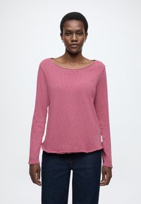 Garām piedurknēm tops mīkstā rozā audumā ar plašu izgriezumu, vaļēju piegriezumu un apaļu apakšu, kombinēts ar tumši ziliem džinsiem.