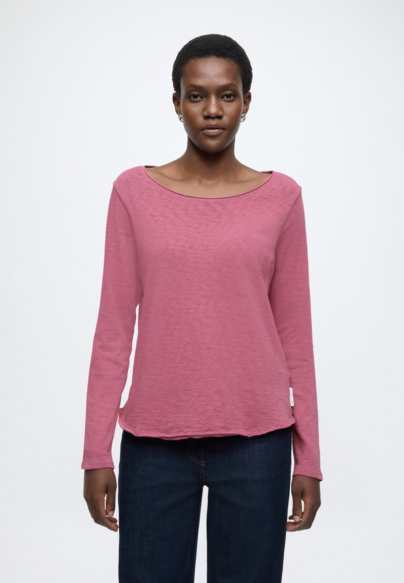 Garām piedurknēm tops mīkstā rozā audumā ar plašu izgriezumu, vaļēju piegriezumu un apaļu apakšu, kombinēts ar tumši ziliem džinsiem.