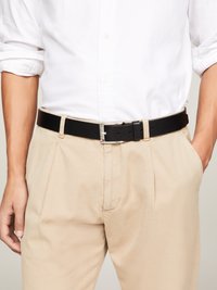 Ceinture en cuir noir avec une boucle en métal argenté, portée avec un pantalon beige, présentant une texture lisse et un design classique.