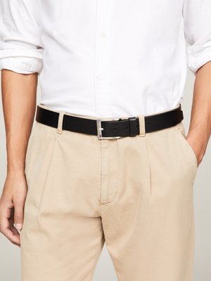 Cintura in pelle nera con fibbia in metallo argentato, indossata con pantaloni beige, caratterizzata da una texture liscia e un design classico.
