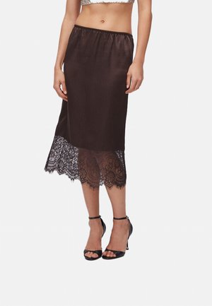 Donna che indossa una gonna midi in satin nero con rifinitura in pizzo sull'orlo e sandali neri con cinturino alla caviglia e tacco alto, posizionata davanti a uno sfondo bianco.