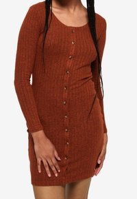 Femme portant une robe boutonnée à manches longues, en côtes, de couleur rouille, avec des boutons sur le devant.