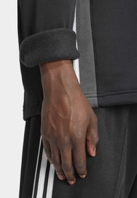 Černý sportovní dlouhý rukáv s měkkým černým fleecovým manžetou; zdobený šedými panely a bílými pruhy po stranách.