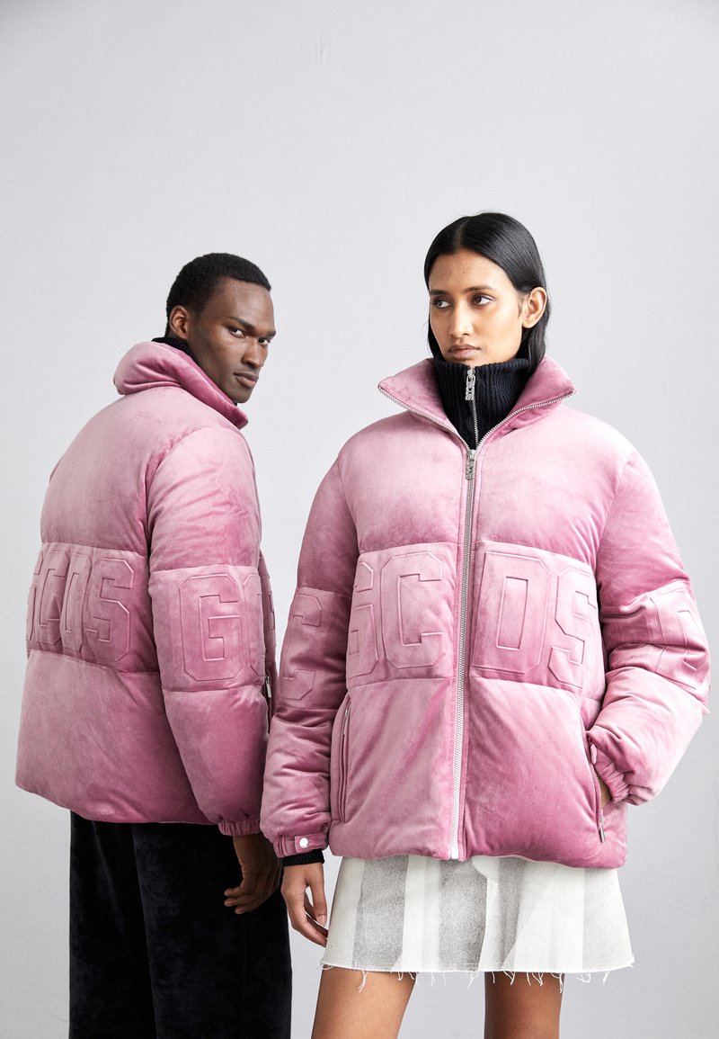 I piumini rosa presentano un design trapuntato con lettere audaci sul petto, colletto alto e chiusure con zip. Un modello indossa una minigonna.