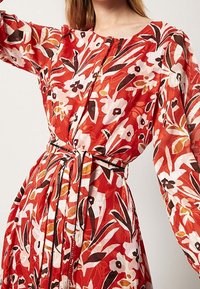 Robe florale rouge à manches longues, avec un motif en blanc et marron. Inclut une ceinture en tissu à la taille.