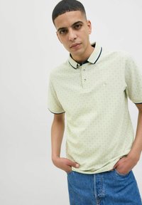 Svetlozelené polo tričko s geometrickým vzorom, so tmavomodrým golierom a zapínaním na tri gombíky. Nosí sa s modrými džínovými šortkami.