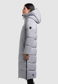 Long, puffy gray coat with a hood, featuring elastic drawstrings, side pockets, and a logo patch on the sleeve. Smooth texture and zippers.