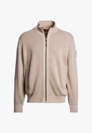 Veste beige zippée avec un col montant, une texture côtelée et des poignets élastiques. Comprend des poches latérales et un logo discret sur la manche.