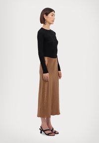 Svart ribbet langermet cardigan med knapper, kombinert med en blank bronsefarget midi-skjørt og svarte stroppsandaler. Glatte teksturer og figursydd design.