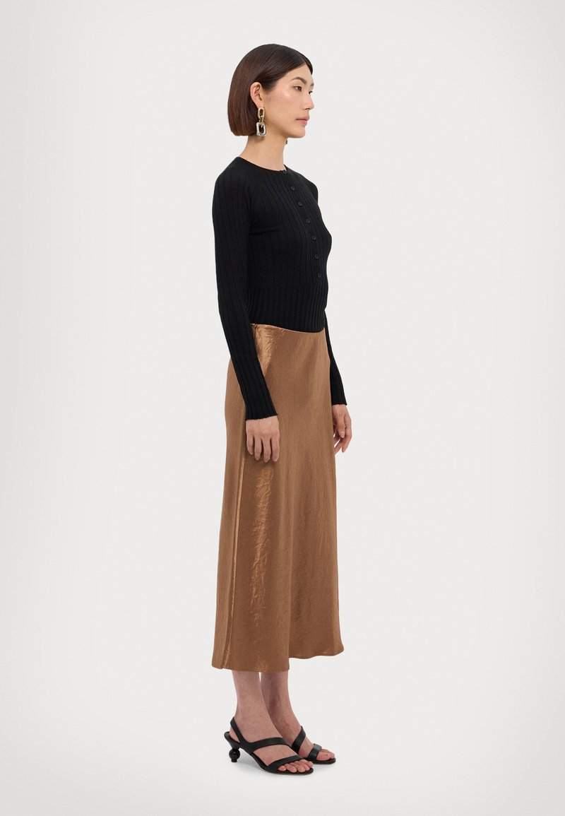 Svart ribbet langermet cardigan med knapper, kombinert med en blank bronsefarget midi-skjørt og svarte stroppsandaler. Glatte teksturer og figursydd design.