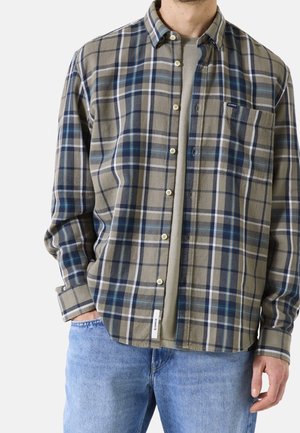 Mies, joka käyttää sinivalkobeige-ruudullista nappipaitaa beigenvärisen t-paidan päällä, siniset farkut, kädet taskuissa ja niiden vieressä.