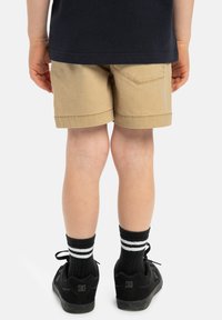 Bruna bomullsshorts med uppvikt nederkant och en lapplomma, bärs med svarta skor och randiga strumpor. Tydlig fokus på shortsens bakvy.