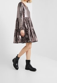 Robe rose doré scintillante avec manches longues et design en couches, associée à des bottines en cuir noir avec boucles et semelles texturées.