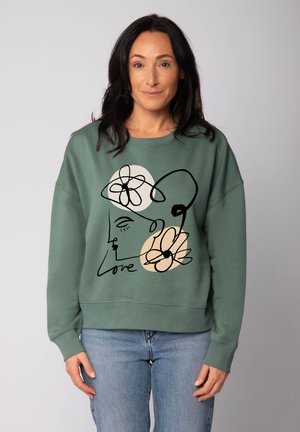 Sweatshirt vert avec un dessin en ligne noire d'un visage et de fleurs, agrémenté d'une touche beige. Texture douce et coupe décontractée.