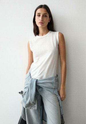 Femme aux cheveux longs et foncés portant un haut blanc sans manches et un jean bleu clair avec une veste en denim nouée autour de la taille, debout contre un mur blanc.