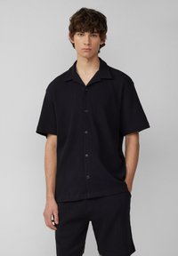Chemise à manches courtes noire à boutons avec un motif texturé, dotée d'un col camp et de boutons noirs, assortie à un short noir.