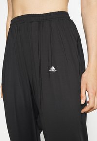 Svarta träningsbyxor med elastisk midja, med vertikala veck och en liten vit Adidas-logga på vänster höft.