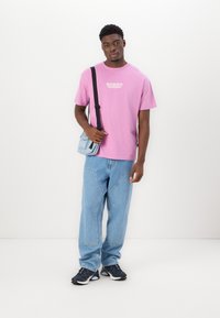 T-shirt en coton rose avec logo, associé à un jean en denim bleu clair à coupe décontractée et des baskets sombres. Petit sac bandoulière bleu.