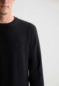 Schwarzer Strickpullover mit rundem Halsausschnitt, strukturiertem Stoff und dezentem Logodetail auf der Brust.