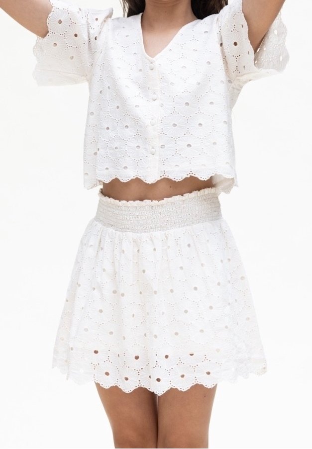 Femme portant une tenue deux pièces en dentelle blanche à œillets, avec un haut court boutonné et une jupe taille haute avec des bords festonnés.
