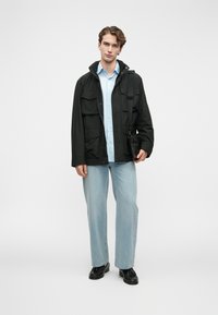 Nuori mies seisoo valkoista taustaa vasten, päällään musta takki, vaaleansininen paita, vaaleat farkut, mustat kengät ja kantaa mustaa olkalaukku. 