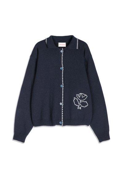 Cardigan bleu marine boutonné avec une bordure triangulaire blanche le long de la ligne de boutons et du col, et motif de fleur brodée blanche sur le bas devant.