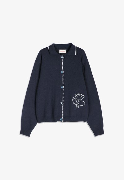Cardigan bleu marine boutonné avec une bordure triangulaire blanche le long de la ligne de boutons et du col, et motif de fleur brodée blanche sur le bas devant.